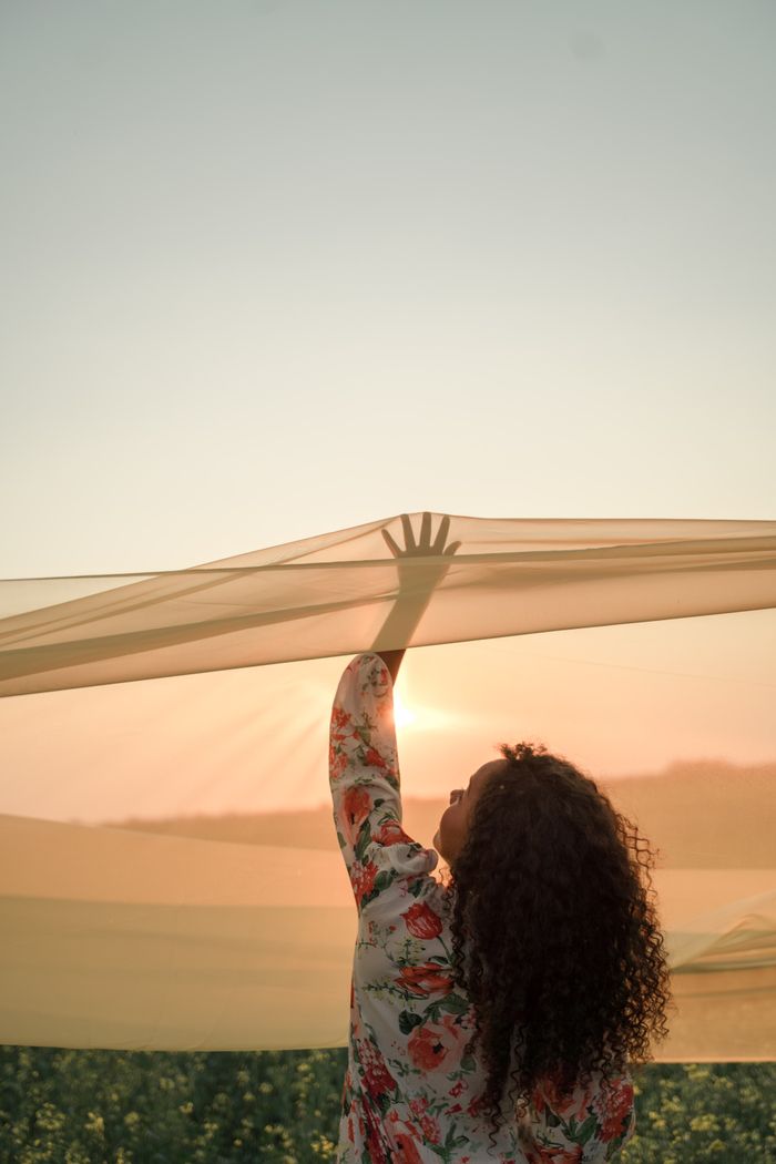A woman stands in a field at sunset, surrounded by flowing fabric, evoking serenity and grace.